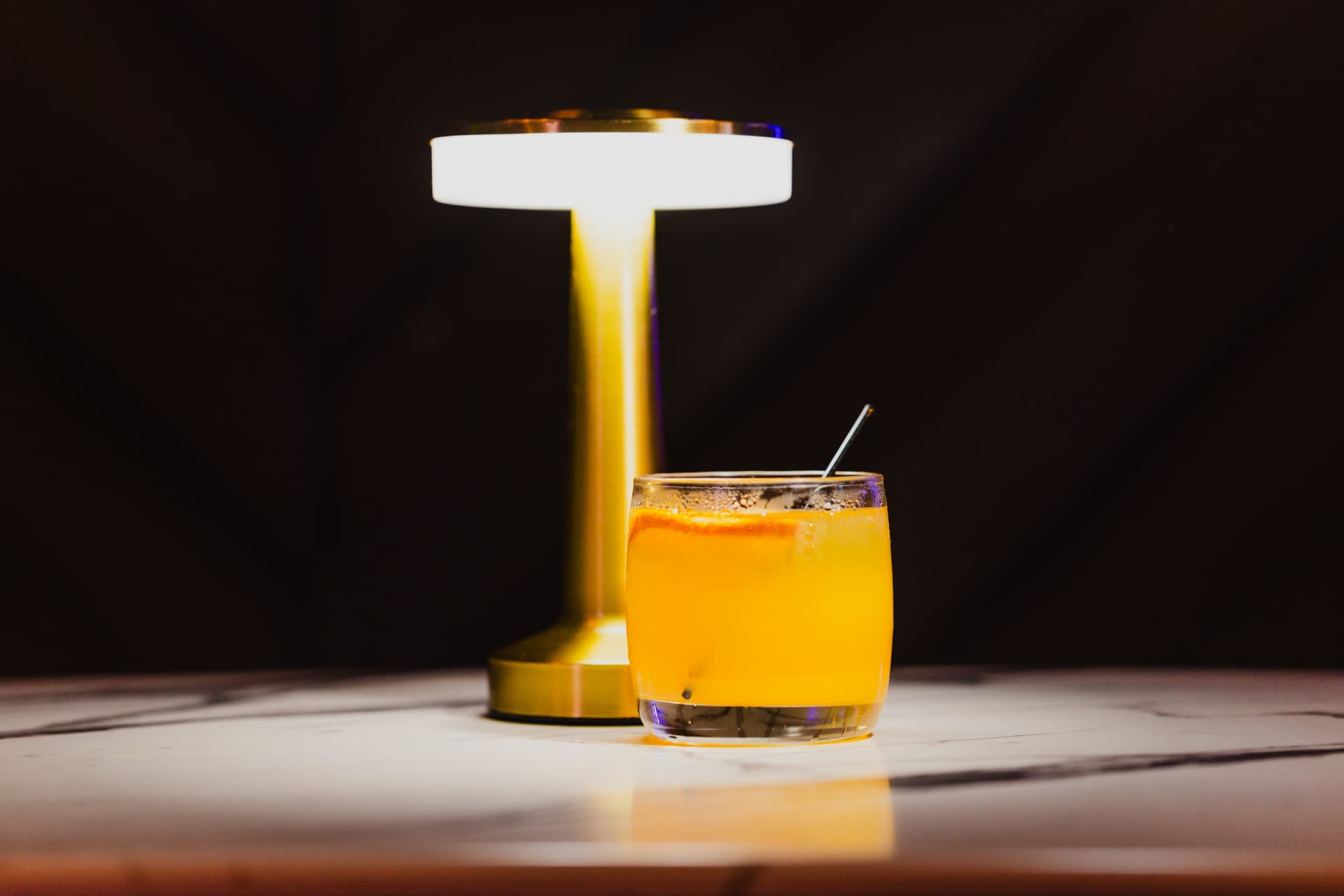 A citrus cocktail glowing beside a gold table lamp at District385